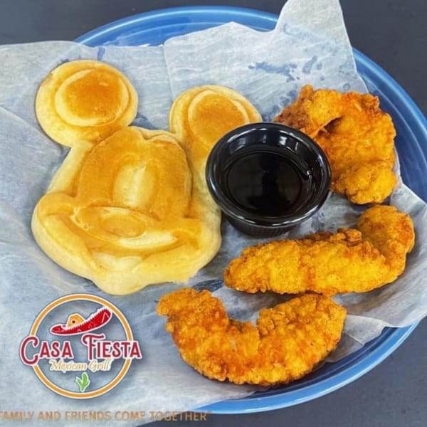 Mickey Mouse-shaped waffle with chicken tenders and a small cup of syrup on a blue plate.