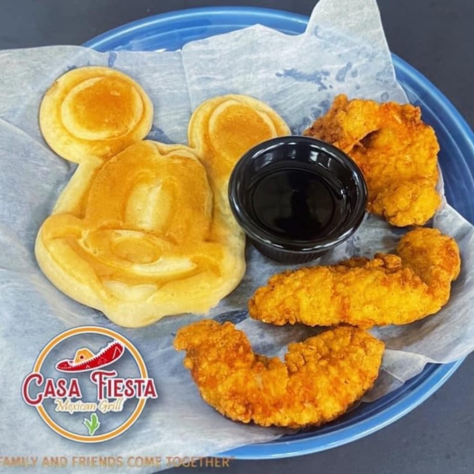 Mickey Mouse-shaped waffle with chicken tenders and a small cup of syrup on a blue plate.
